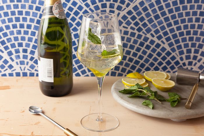 A wine glass filled with a clear drink, lemon slices, and mint leaves sits on a table next to a bottle, a spoon, and a round tray with more lemon slices and mint leaves. Blue and white mosaic tile background.