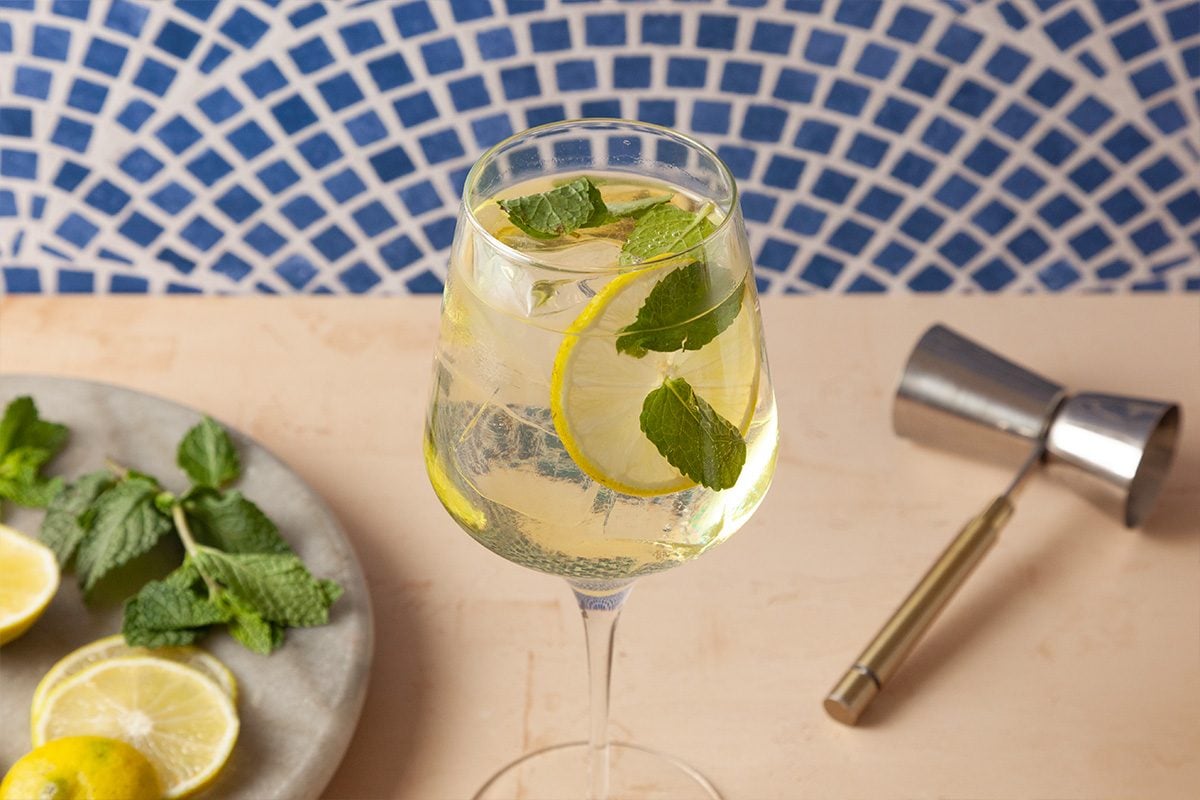 A glass of sparkling drink garnished with lemon slices and mint leaves sits on a light surface, with a plate of mint and lemon and a metal jigger nearby; a blue mosaic tile background is visible.