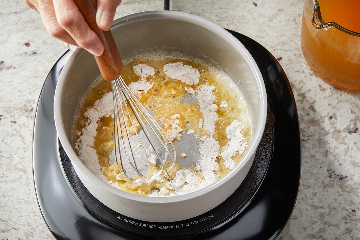 A hand uses a whisk to mix flour and melted butter in a saucepan on a stovetop, beginning to make a roux.
