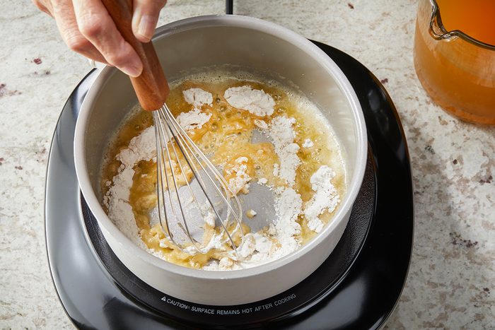 A hand uses a whisk to mix flour and melted butter in a saucepan on a stovetop, beginning to make a roux.