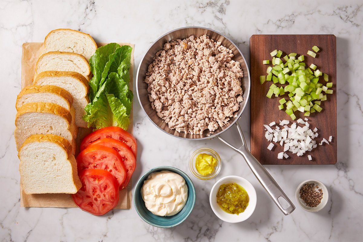 Sliced white bread, lettuce, tomato, mayonnaise, mustard, relish, ground meat in a pan, and chopped celery and onion on a cutting board arranged on a marble surface.