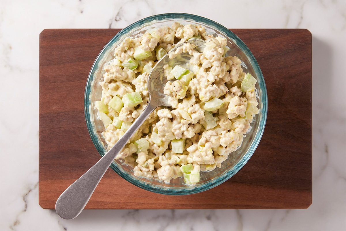 A glass bowl filled with chicken salad containing diced celery and a creamy dressing sits on a wooden cutting board; a metal spoon rests inside the bowl.