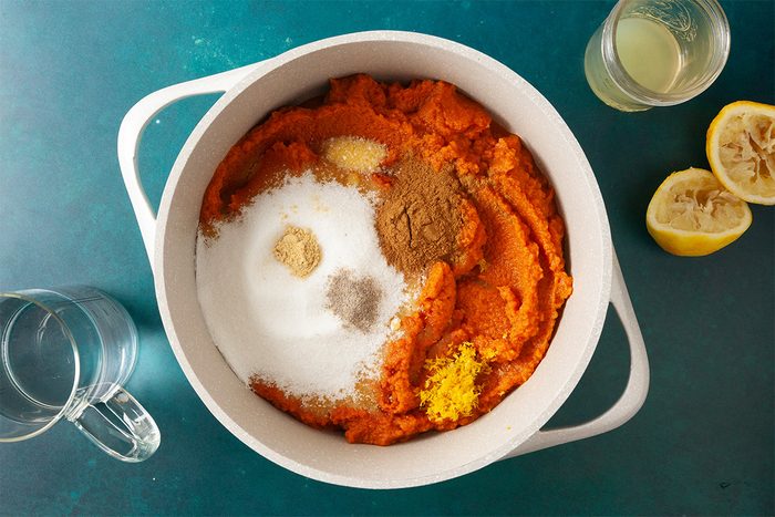 A white pot filled with pumpkin puree, sugar, spices, and lemon zest sits on a teal surface. Nearby are a glass of water, a glass of juice, and two squeezed lemon halves.