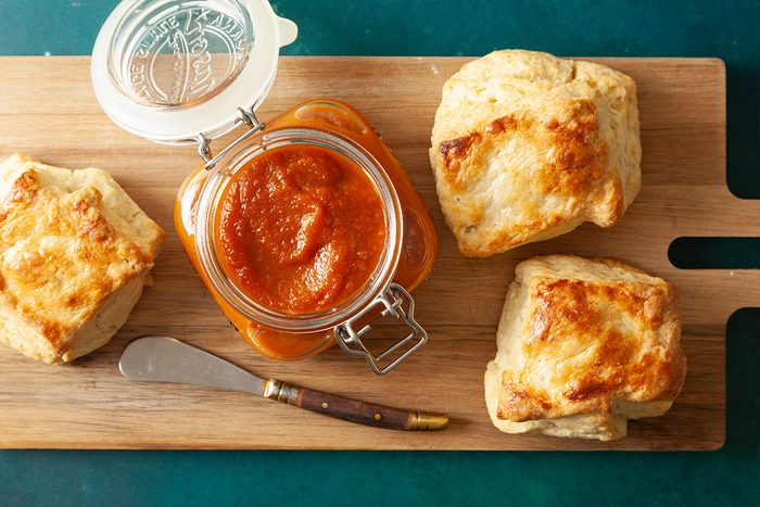 Three golden, flaky biscuits sit on a wooden board next to an open glass jar filled with red tomato sauce. A butter knife with a dark handle rests beside the jar.