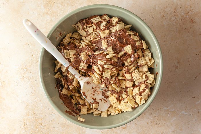 Overhead shot of a green bowl filled with Chex cereal coated in chocolate, with a white spatula resting inside; placed on a light beige countertop
