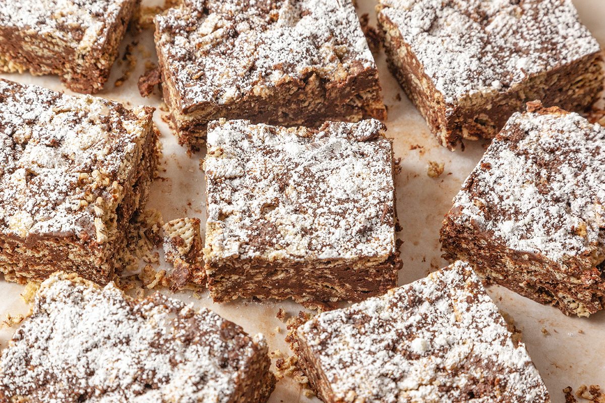 Close-up shot of chocolate Puppy Chow Bars cut into squares and dusted with powdered sugar; arranged on a parchment-lined surface; The texture of the cereal and chocolate mixture is visible on the sides