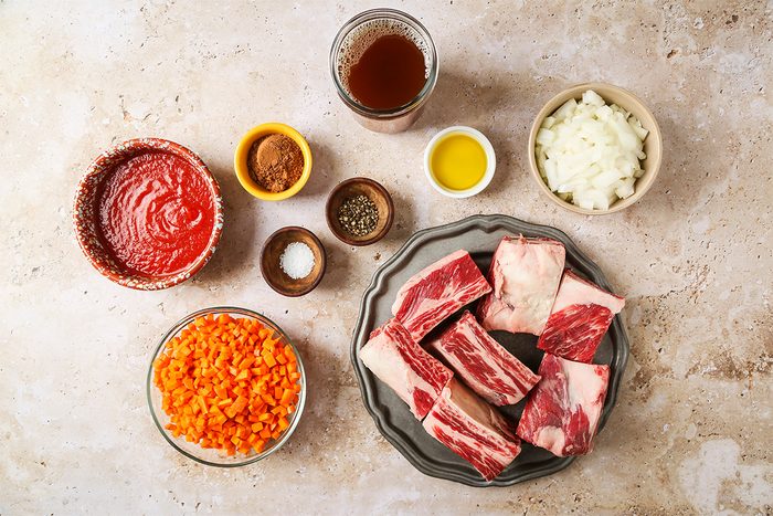 A top-down view of ingredients for a beef dish, including raw beef short ribs, chopped carrots, diced onions, tomato sauce, olive oil, broth, and small bowls of spices arranged on a light surface.
