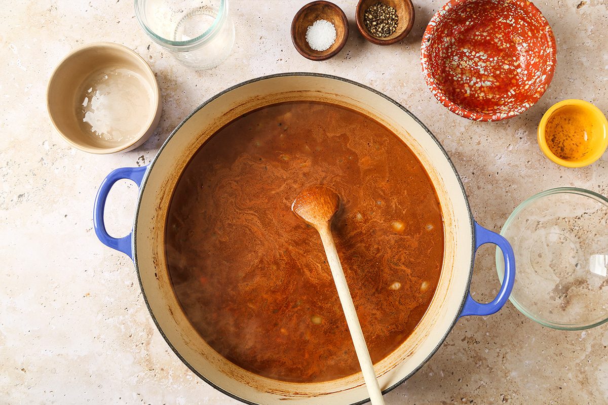 A blue pot filled with simmering brown soup and a wooden spoon, surrounded by small bowls containing seasonings and ingredients, all on a light countertop.