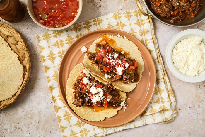 Two tacos filled with seasoned beef, diced tomatoes, and crumbled cheese sit on a pink plate. Surrounding the plate are bowls of salsa, cheese, and tortillas on a patterned cloth background.
