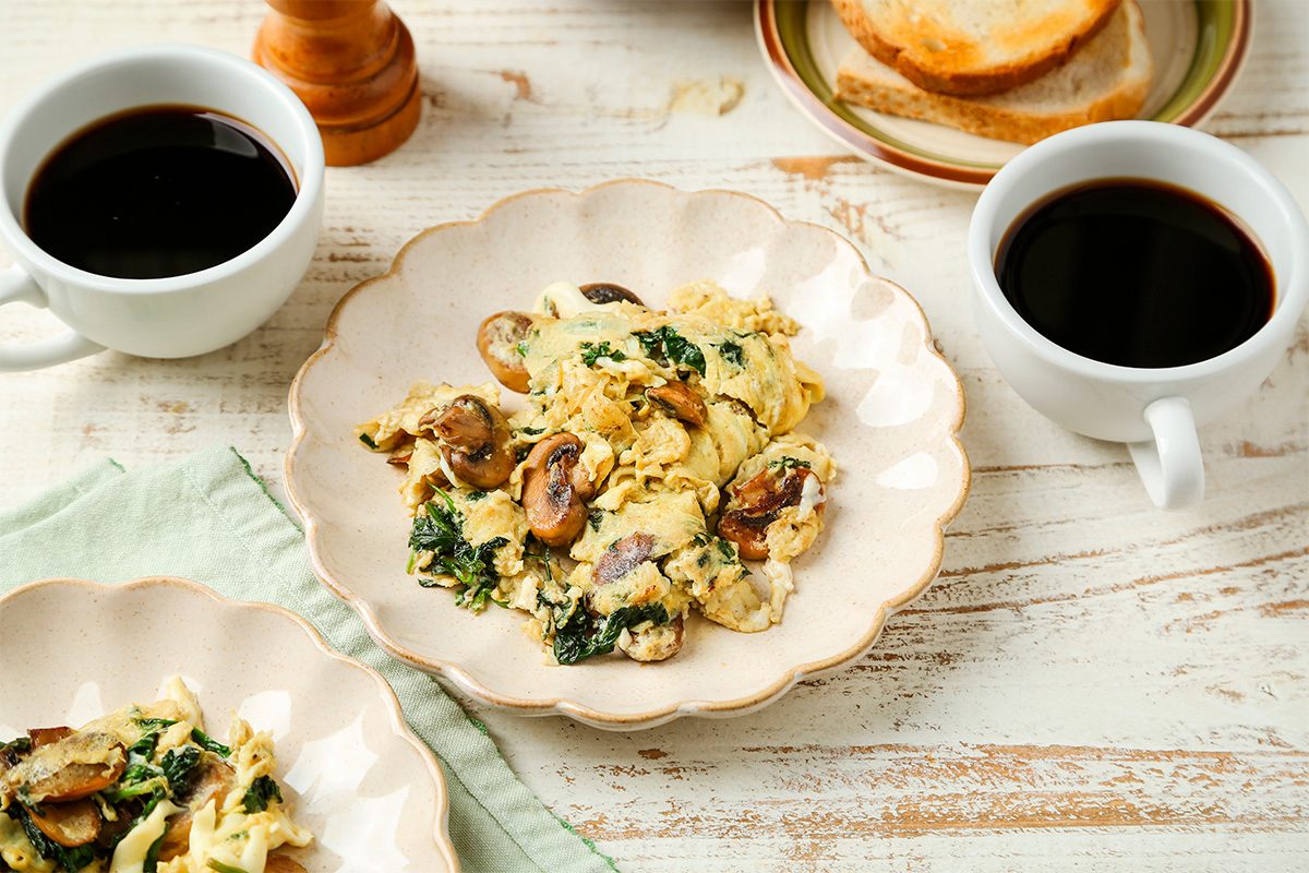 A plate of scrambled eggs with spinach and mushrooms is served on a light-colored table, accompanied by two cups of black coffee and a plate of toasted bread slices in the background.