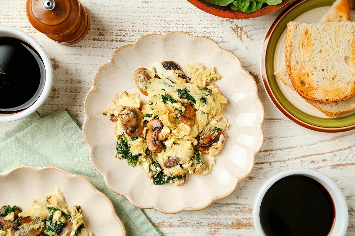 A plate of scrambled eggs with mushrooms and spinach on a cream-colored plate, next to slices of toasted bread, two cups of coffee, and a pepper grinder on a rustic white table.