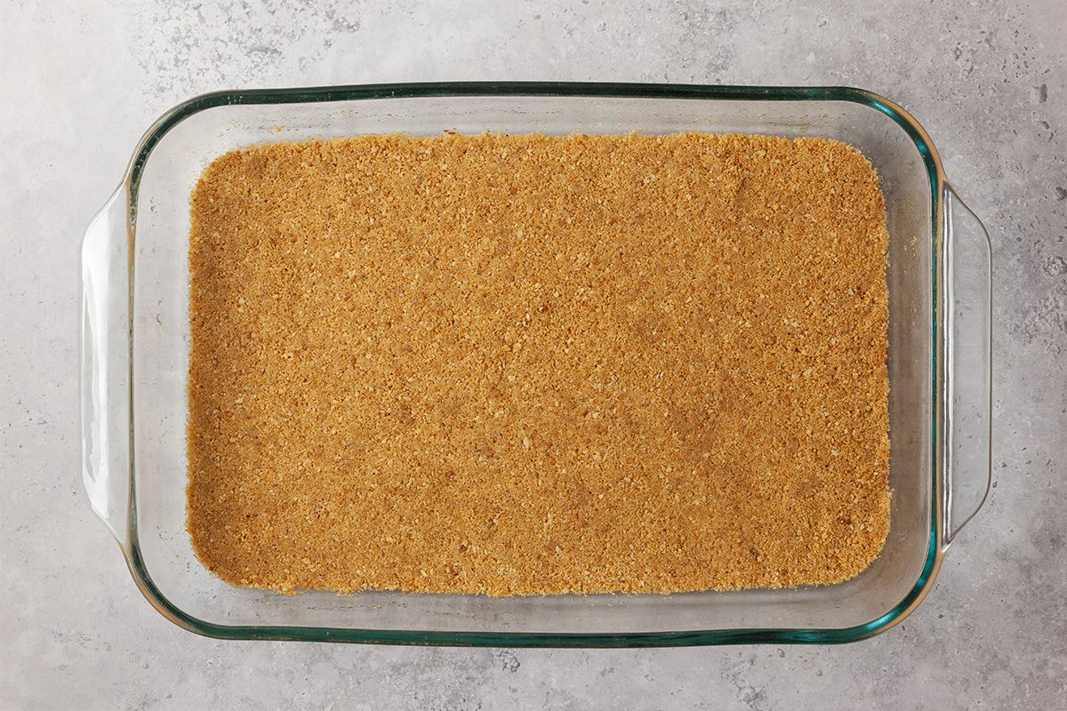 A glass baking dish filled with an even layer of graham cracker crust, pressed down and ready for baking, sits on a light gray countertop.