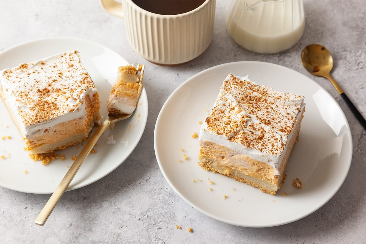Two plates each with a square slice of layered cake topped with whipped cream and cinnamon, one with a fork cutting a bite. A cup and a small jug, possibly with milk, are in the background.