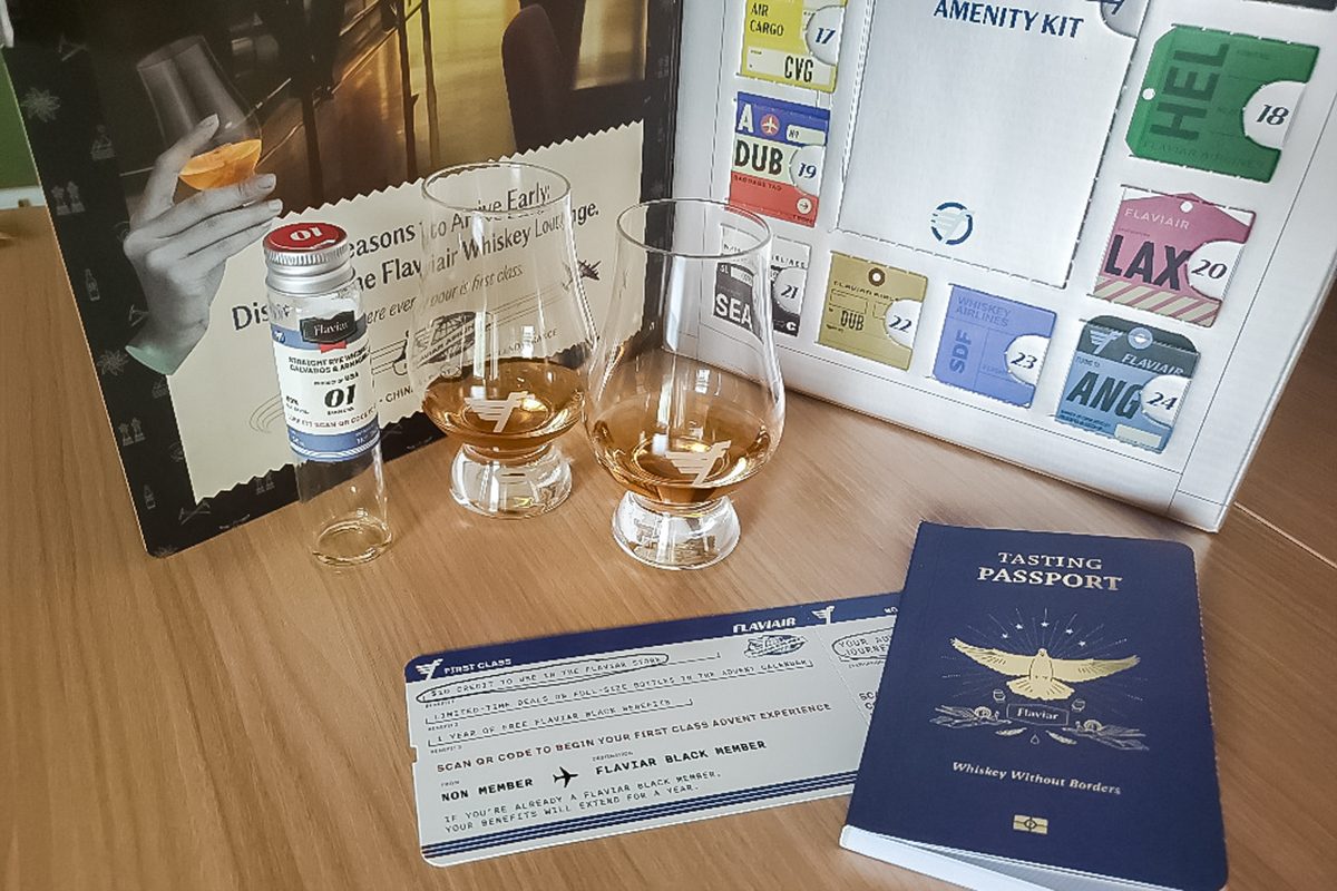 A display on a wooden table features two whiskey glasses, a small whiskey bottle, a blue travel passport, a mock boarding pass, and a colorful board with airport codes and an amenity kit card.