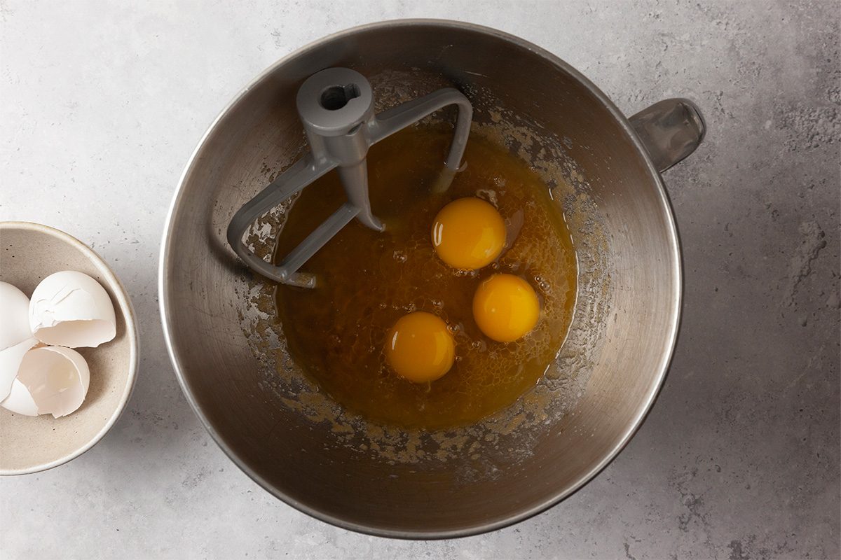 A metal mixing bowl with three raw eggs and sugar inside, fitted with a paddle attachment, sits on a light gray countertop. A small bowl with empty eggshells is beside it.