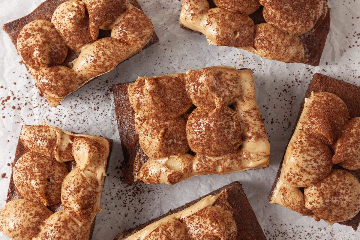 Six rectangular tiramisu brownies with creamy, swirled topping and a dusting of cocoa powder, arranged on a piece of parchment paper.