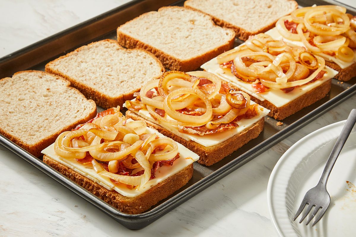 3/4th shot of Slices of bread on a baking sheet, some topped with cheese, crispy bacon, and caramelized onions, ready to be assembled into sandwiches; A white plate with a fork is nearby