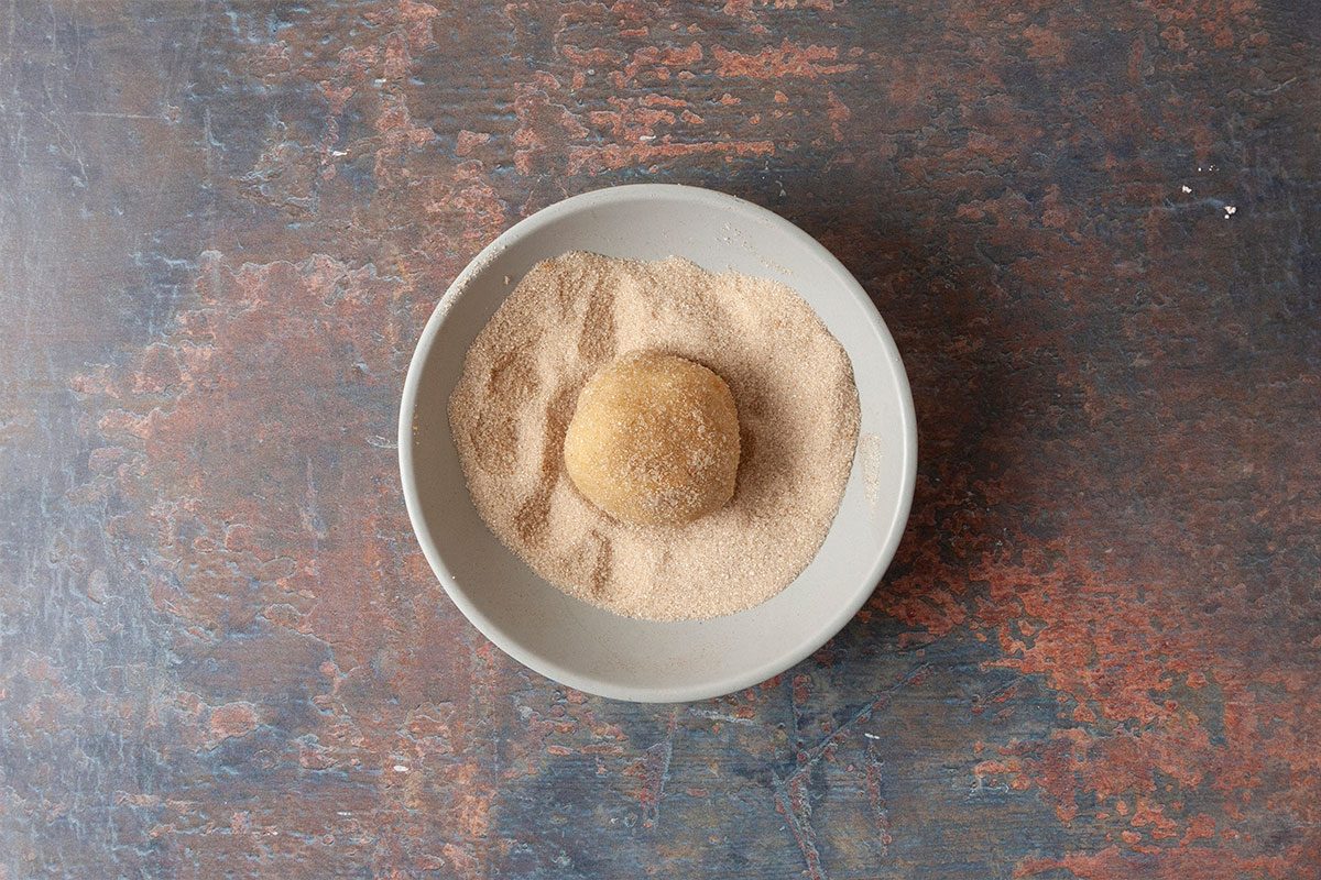 Overhead shot of a round dough ball in a shallow bowl of cinnamon sugar, ready to be coated, set on a textured brown and blue surface