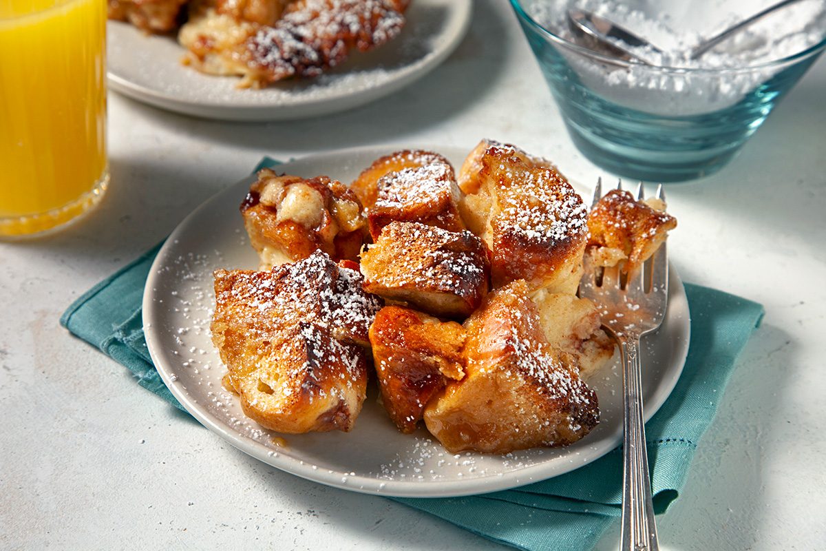 A plate of golden brown, baked French toast pieces dusted with powdered sugar sits on a teal napkin, with a fork beside it. In the background are a glass of orange juice and a bowl of powdered sugar.