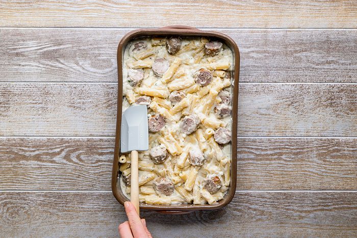 A hand holding a spatula is spreading a creamy baked pasta dish with sausage slices in a rectangular baking dish on a wooden surface.