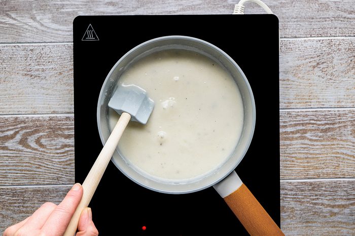 A hand holding a spatula stirs a pot of white sauce on an electric stovetop with a wooden surface underneath.