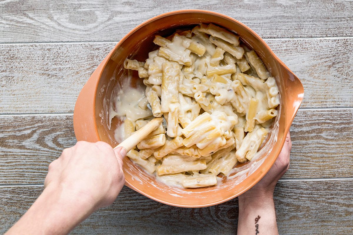 A person stirs cooked pasta with creamy white sauce in an orange mixing bowl, placed on a light wooden surface. One hand holds the bowl while the other stirs with a spatula.