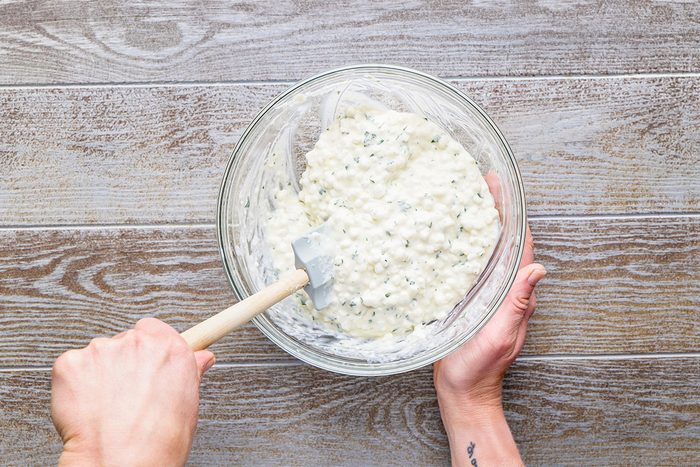 A person holding a glass bowl filled with a creamy, chunky mixture and stirring it with a white spatula, on a wooden surface.