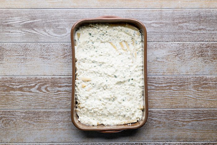 A rectangular baking dish filled with an unbaked lasagna, topped with a thick layer of creamy white cheese mixture, sits on a wooden surface.