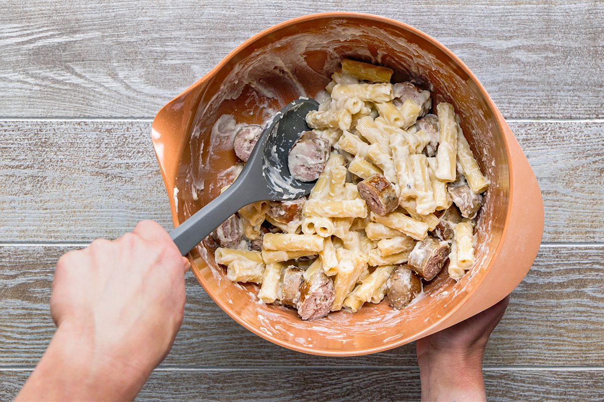 A person stirs creamy pasta with sausage pieces in a large orange mixing bowl using a gray spatula, on a wooden surface.