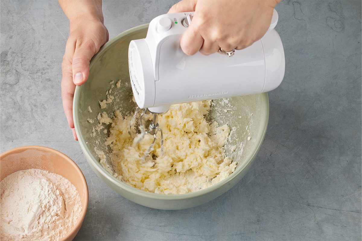 Overhead shot of a person using a white hand mixer to blend ingredients in a light green bowl, with a peach-colored bowl of flour nearby on a gray countertop;