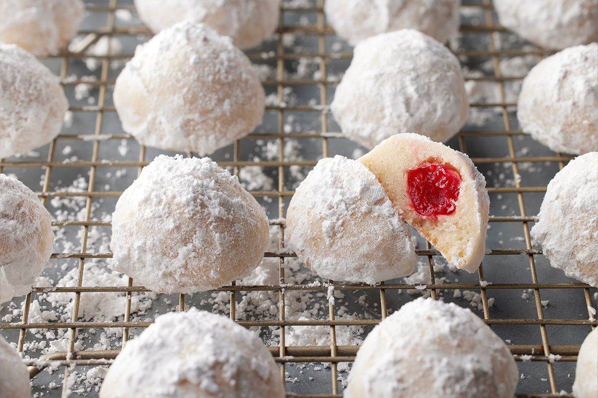 3/4 angle view shot of cherry snowball cookies dusted with powdered sugar resting on a cooling rack, with one revealing a vibrant red cherry filling inside;
