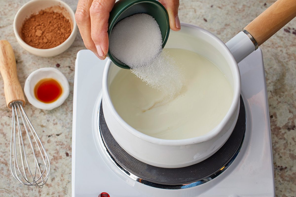 Overhead shot of a hand pouring sugar into a white pot of milk on a stovetop