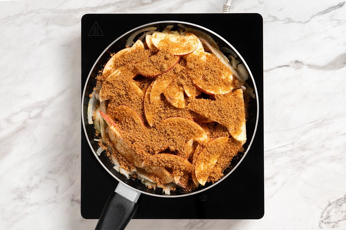 Overhead shot of the same skillet; sprinkle over apples; cover and simmer for 10 minutes; induction; all set on a white marble surface;