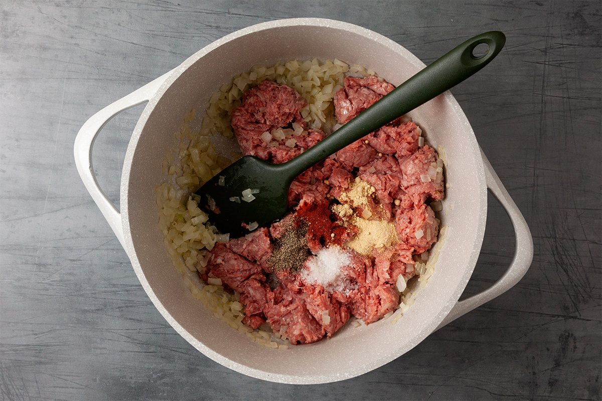 Overhead shot of a white pot holds chopped onions raw ground beef and various spices and a green spatula rests on its edge on a gray surface