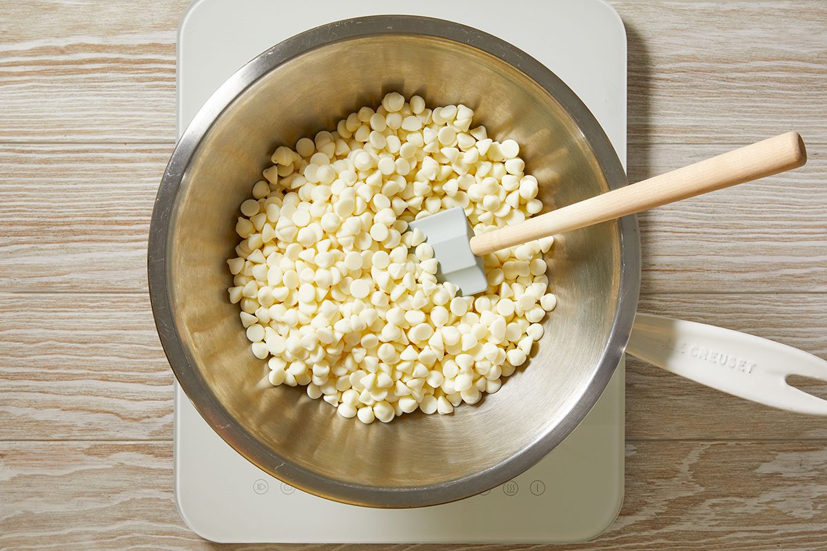 A metal bowl filled with white chocolate chips and a spatula resting inside, placed on a light-colored surface.