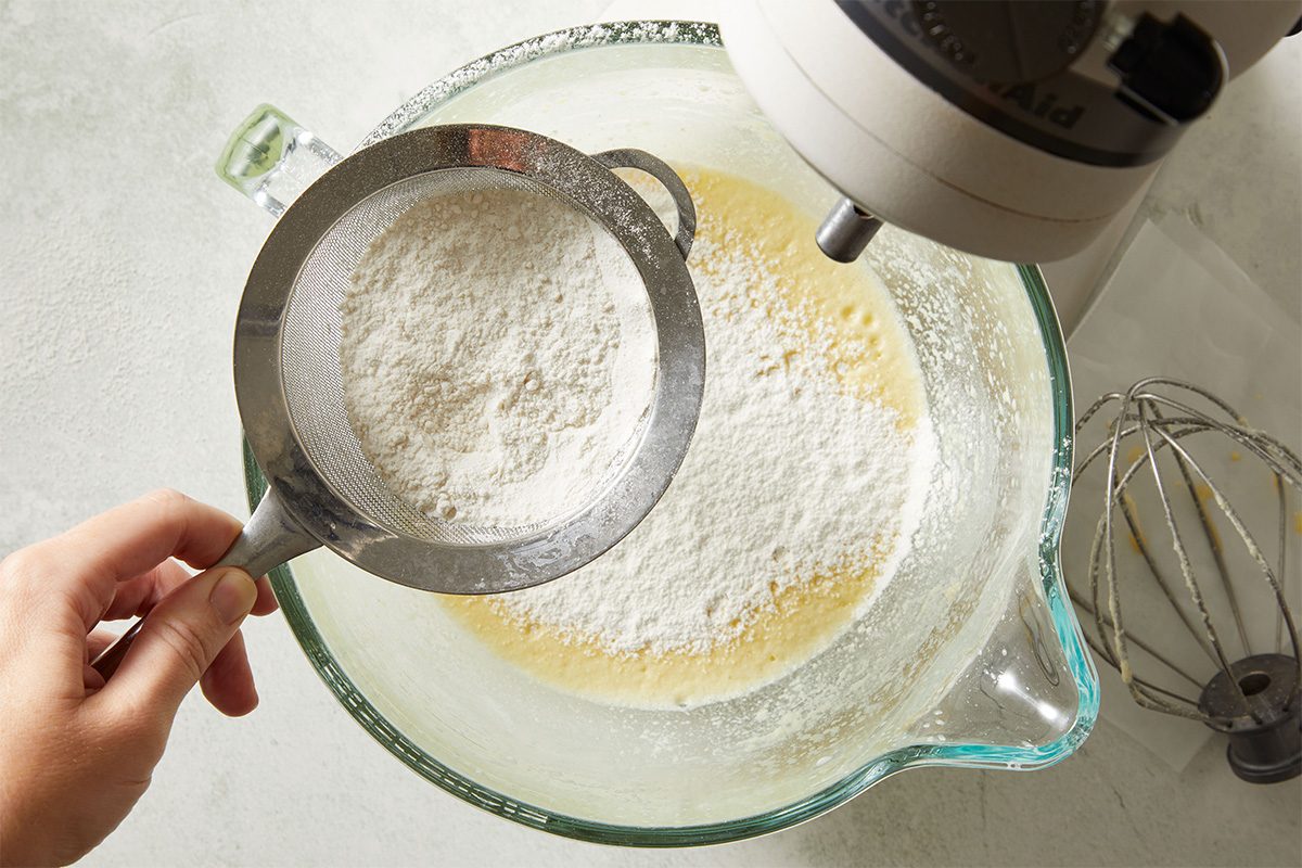 A hand sifts flour into a glass mixing bowl with wet ingredients under a stand mixer, with a whisk attachment and parchment paper nearby on a light-colored surface.