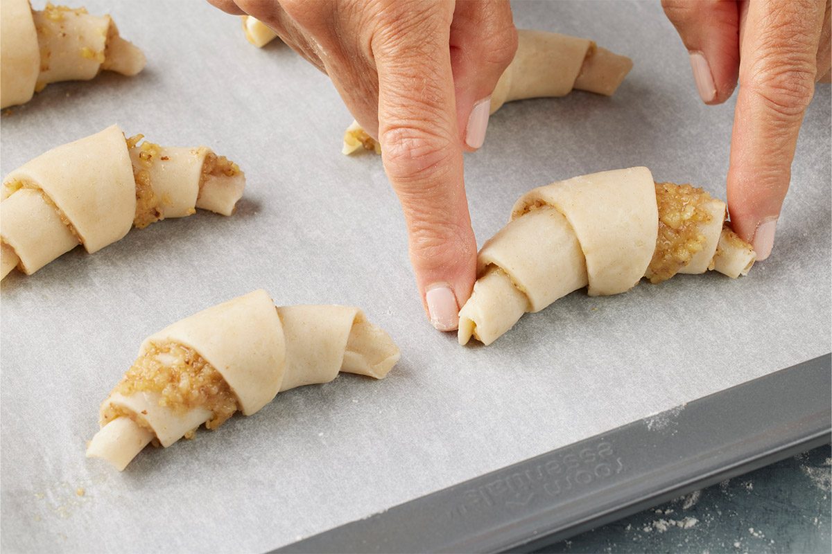 Close-up of hands rolling crescent-shaped pastries filled with a nut mixture on a parchment-lined baking sheet;