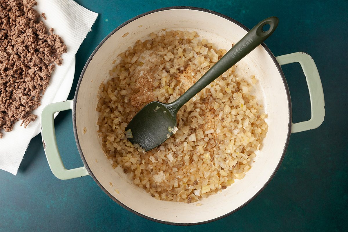 Overhead shot of a pot filled with sautéed chopped onions and a green spatula, resting on a teal surface; cooked ground beef drains on a paper towel nearby