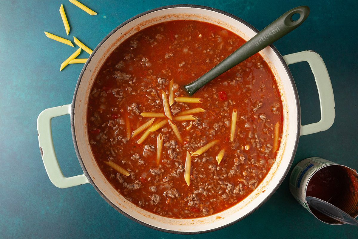 Overhead shot of a large pot of tomato-based meat sauce with uncooked penne pasta scattered on top and around the teal surface; a green-handled spoon rests inside the pot, and an open can sits nearby