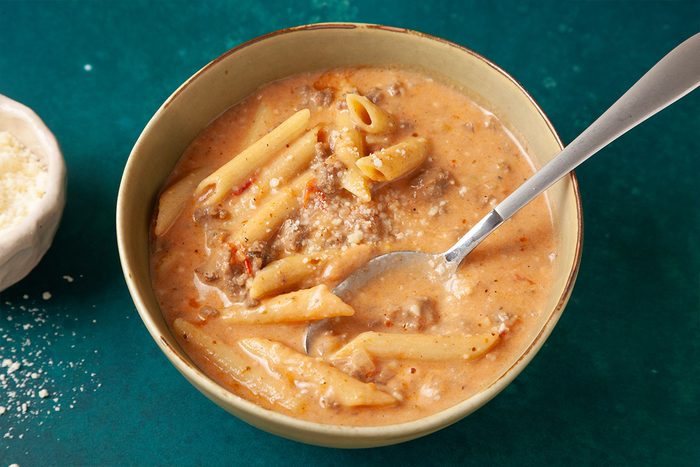 Close shot of a bowl of Pastitsio Soup with penne pasta, ground meat, tomato, and grated cheese; a spoon rests in the bowl, with a small dish of extra cheese nearby