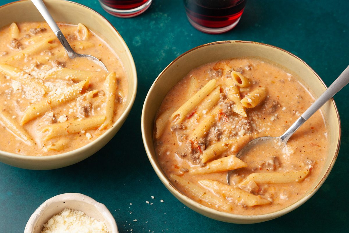 High angle view shot of two bowls of Pastitsio Soup with penne, ground meat, and tomato broth on a green surface; spoons, grated cheese, and red drinks are arranged nearby
