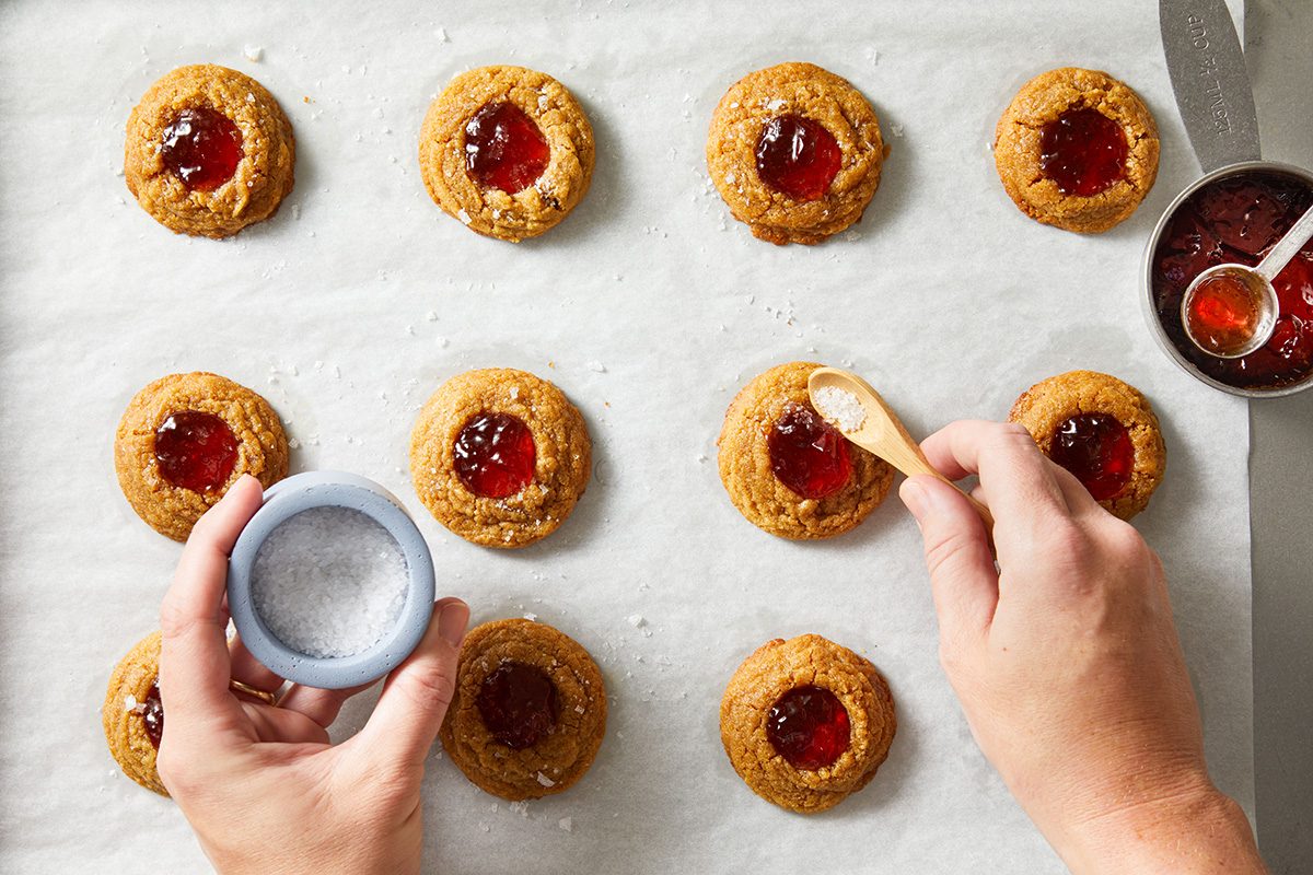 Overhead shot of a hand spooning bright red jelly into the center of peanut butter cookie wells