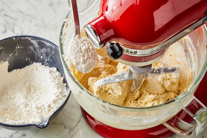 A red stand mixer blends dough in a glass bowl, while a spoon adds flour from a nearby black bowl on a marble countertop.
