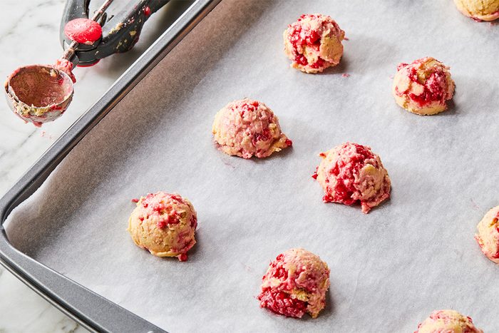 A baking sheet lined with parchment paper holds several unbaked cookie dough balls with red fruit pieces. A cookie scoop with dough residue is resting beside the sheet.