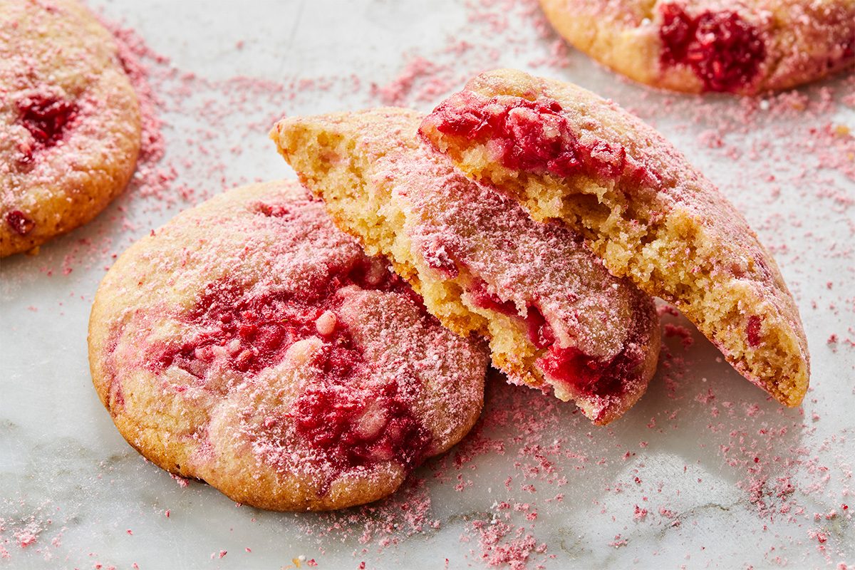 Two strawberry cookies on a white surface, one whole and one broken in half, revealing a soft, crumbly inside with visible strawberry pieces and a dusting of pink sugar on top and around them.