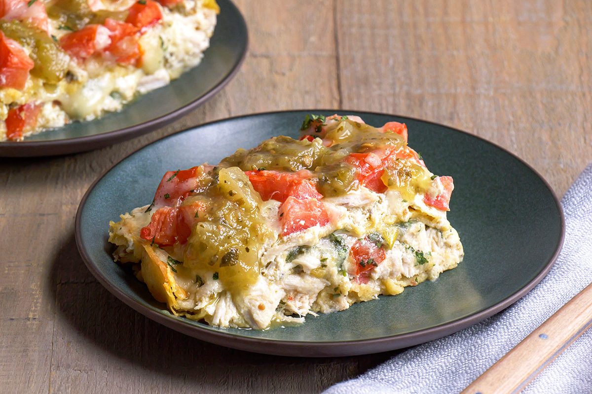 A slice of casserole topped with diced tomatoes and green chiles is served on a dark plate, placed on a wooden table with a napkin and another plate visible in the background.