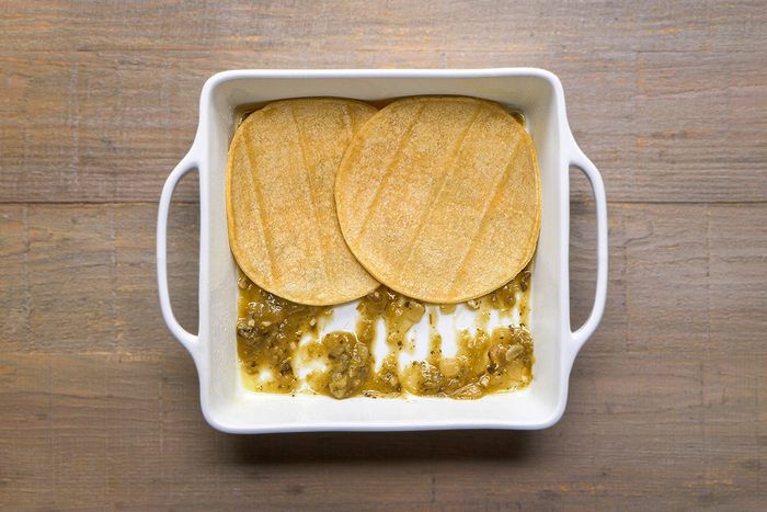 A white square baking dish with handles contains two corn tortillas partially covering a layer of green sauce spread on the bottom, placed on a wooden surface.
