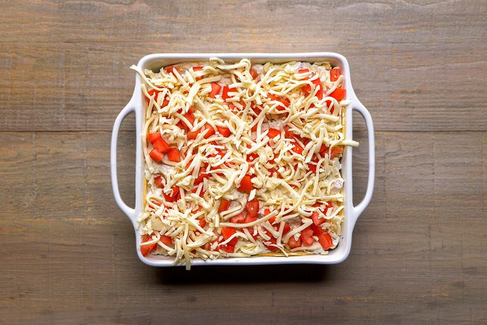 A white square baking dish filled with a casserole topped with shredded cheese and diced red bell peppers, placed on a wooden surface.