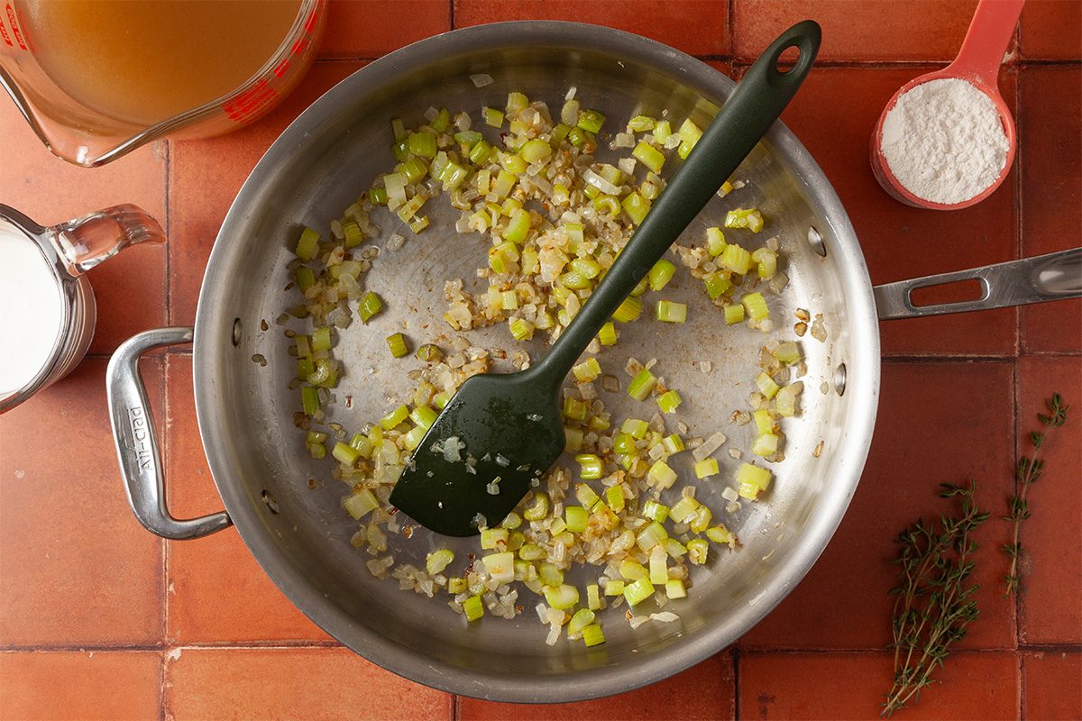 Overhead view of chopped onions and celery being sautéed in a large pan with a black spatula, surrounded by a measuring cup of broth, a cup of milk, a cup of flour, and sprigs of fresh thyme