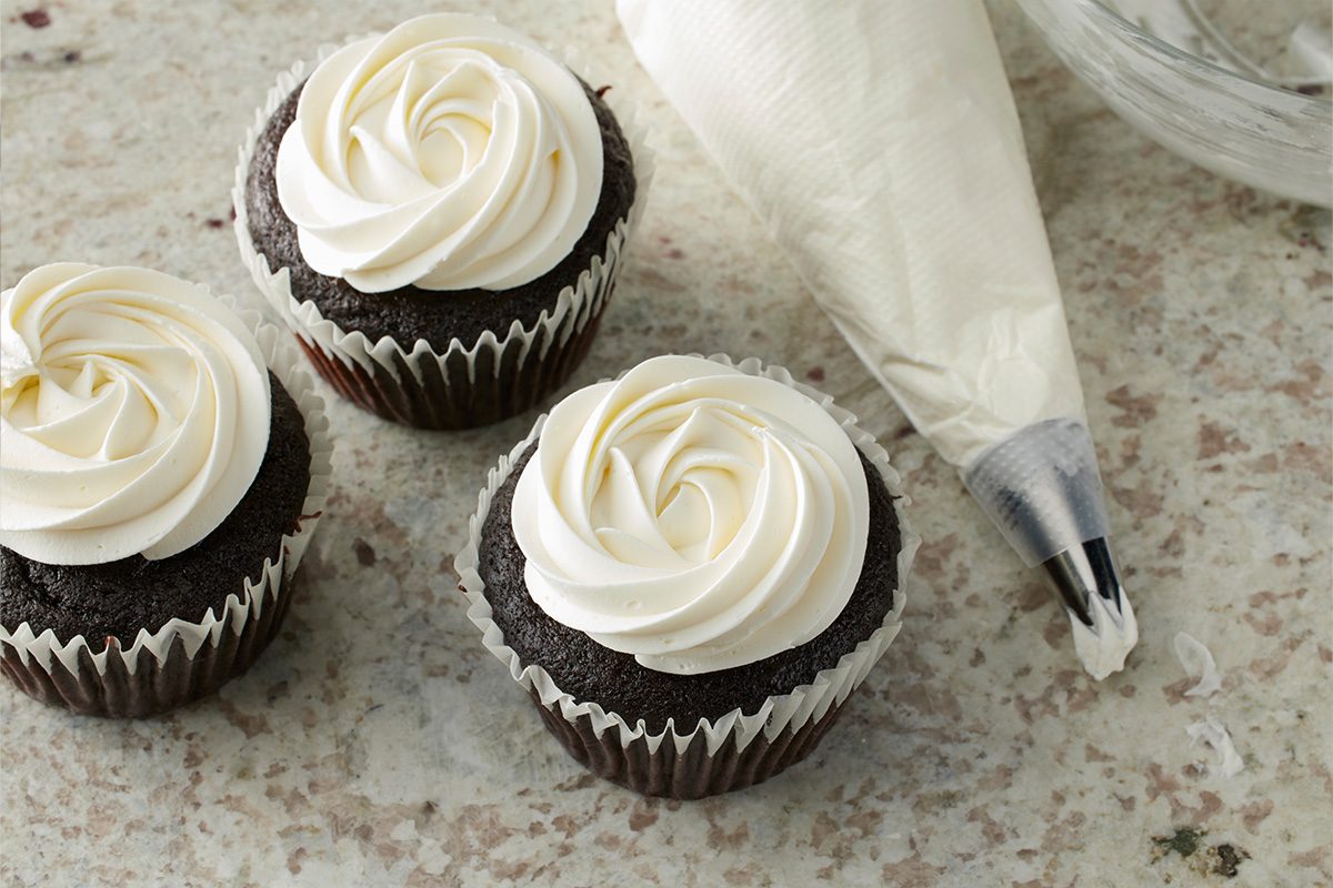 Three chocolate cupcakes with white swirled frosting next to a piping bag filled with frosting, placed on a speckled countertop.